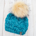 Blue knitted beanie with a large pom-pom on a wooden surface; Nor'easter Knits The Gibbon Beanie Teal Feather 100% Pure Merino Wool Winter Hat with faux fur pom pom
