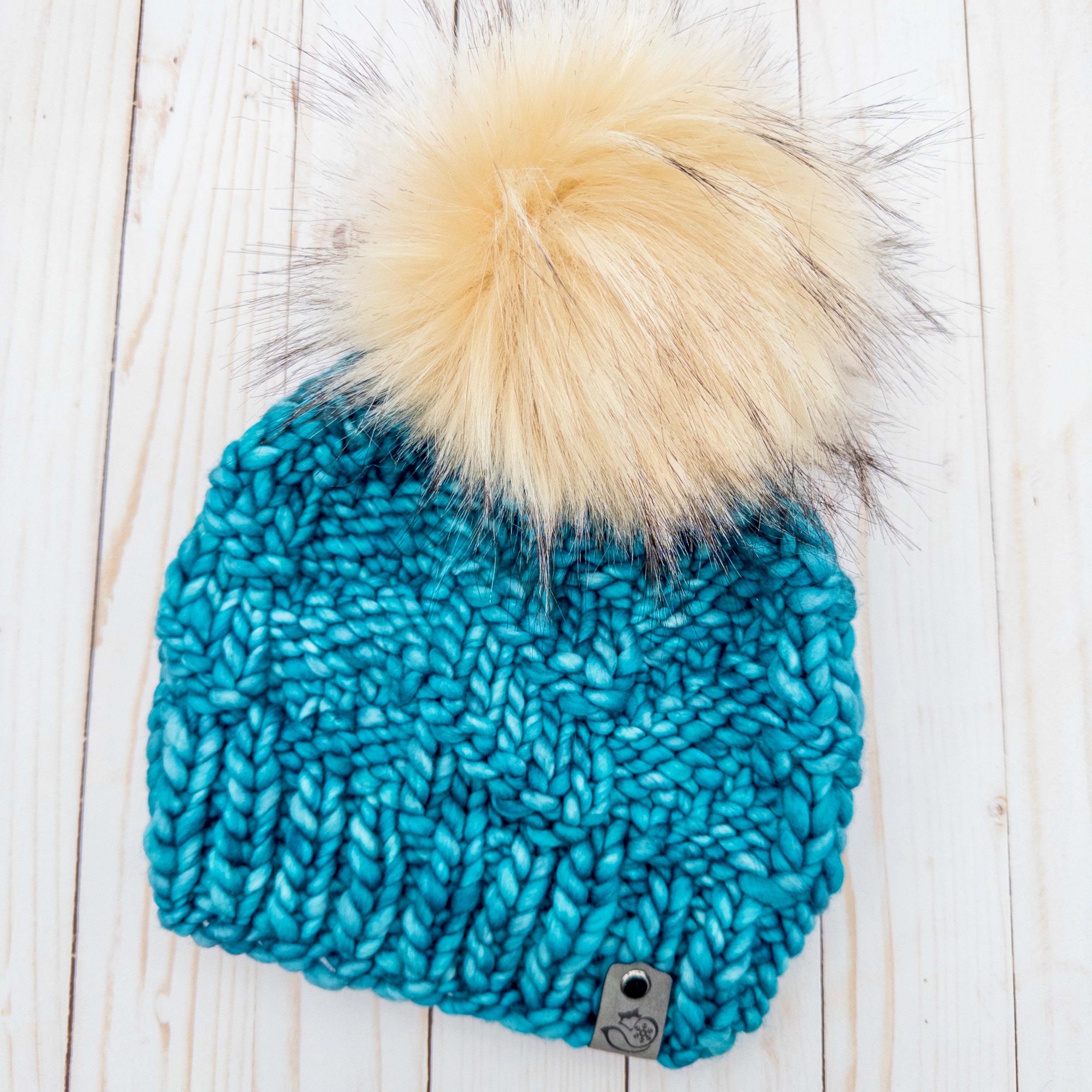 Blue knitted beanie with a large pom-pom on a wooden surface; Nor'easter Knits The Gibbon Beanie Teal Feather 100% Pure Merino Wool Winter Hat with faux fur pom pom