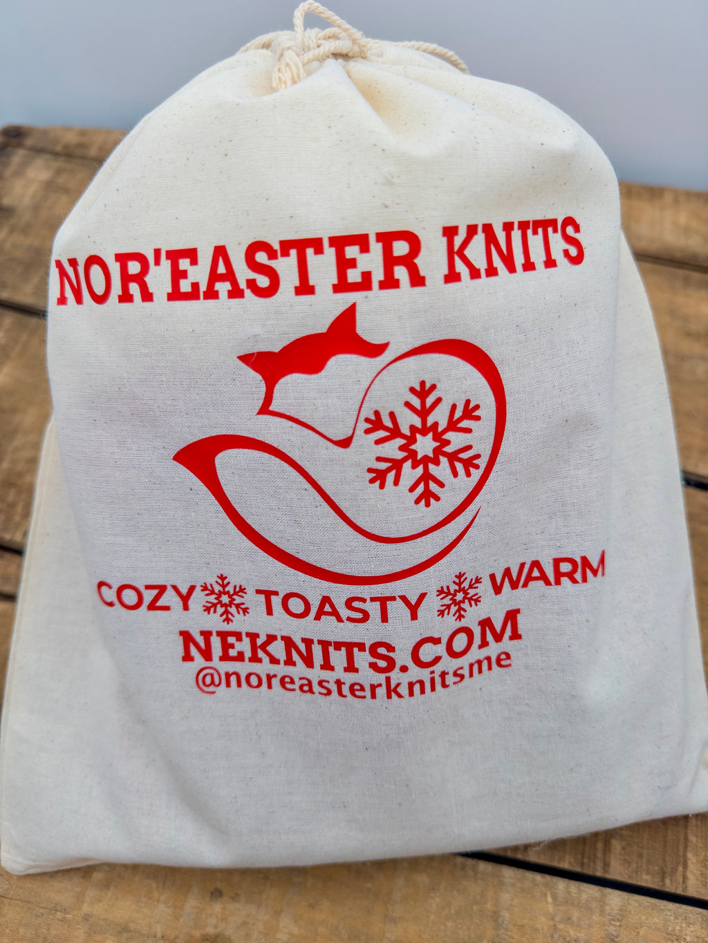 Natural cotton muslin Nor’easter Knits toque tote bag with red logo branding and drawstring closure on rustic wood background