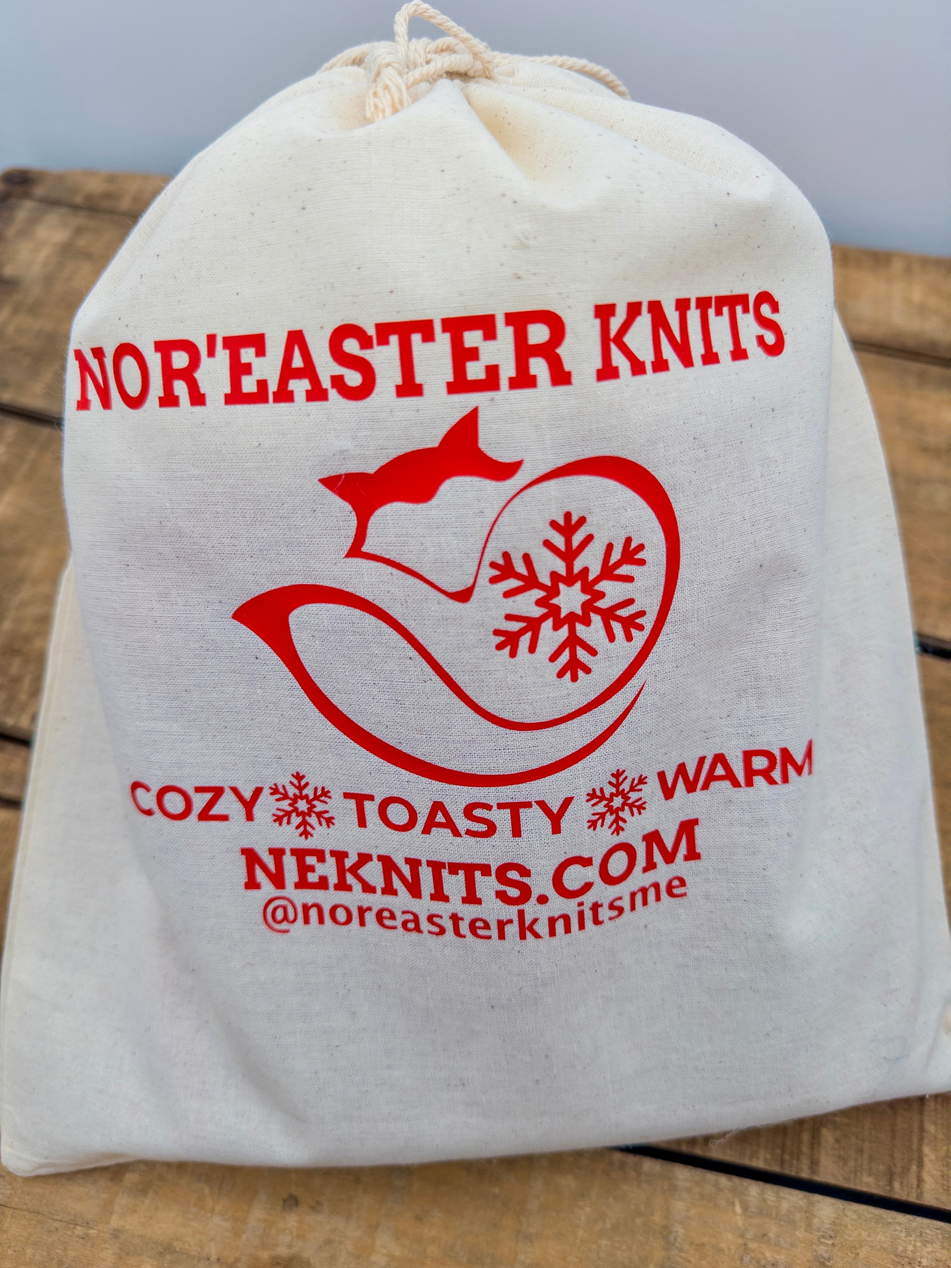 Natural cotton muslin Nor’easter Knits toque tote bag with red logo branding and drawstring closure on rustic wood background